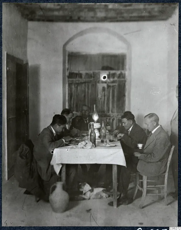Men eating at a table with a checked cloth in a whitewashed room, with a dog on the floor beneath the table