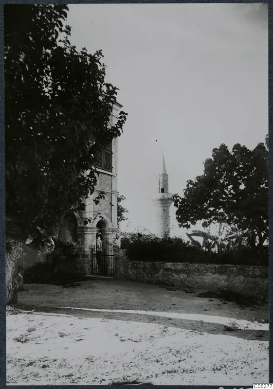Stone building with an arched gateway and iron gate at Lapithos, a large tree beside it and a slender tower behind