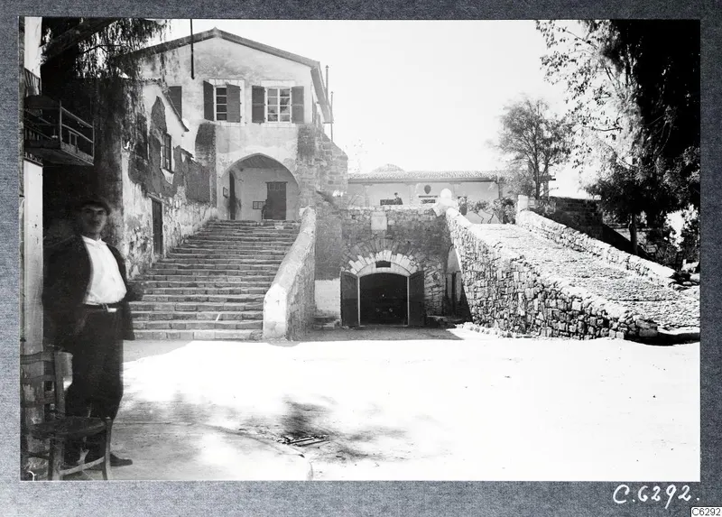Courtyard with a broad stone staircase, an arched doorway and a man standing at left beside a chair