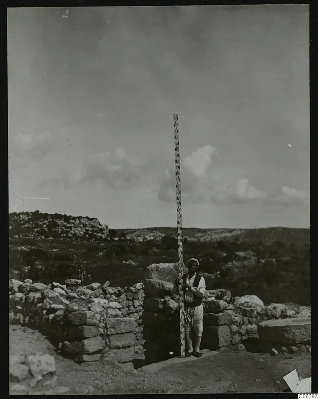 Man holding a tall measuring rod beside stone walls at Nitovikla
