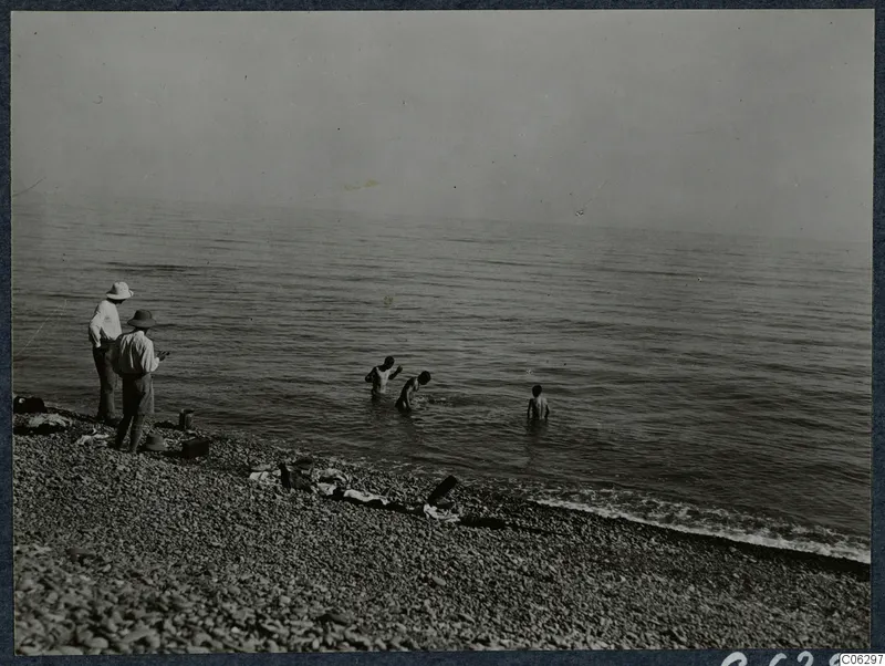 Two men stand on a pebble beach watching three young figures wading in the calm sea