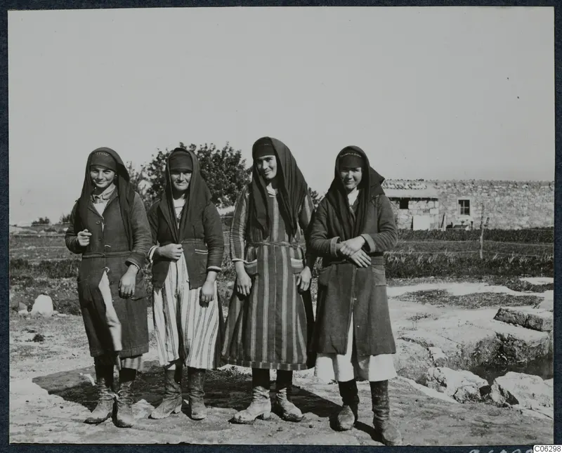 Four young women in headscarves and long coats stand in a row outside, with a stone building behind them, at Vouni.