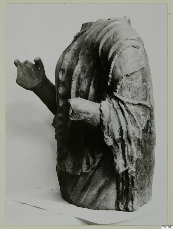 Headless draped figure fragment with one arm raised and the other bent across the body, shown against a pale backdrop