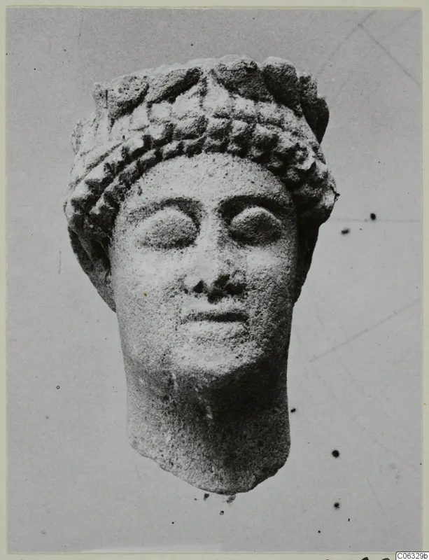 Limestone head of a figure with a banded headdress and wide almond-shaped eyes, photographed against a plain grey background.