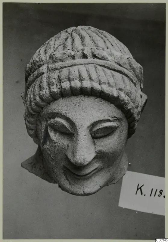 Limestone head of a smiling figure with grooved hair, shown against a dark grey backdrop