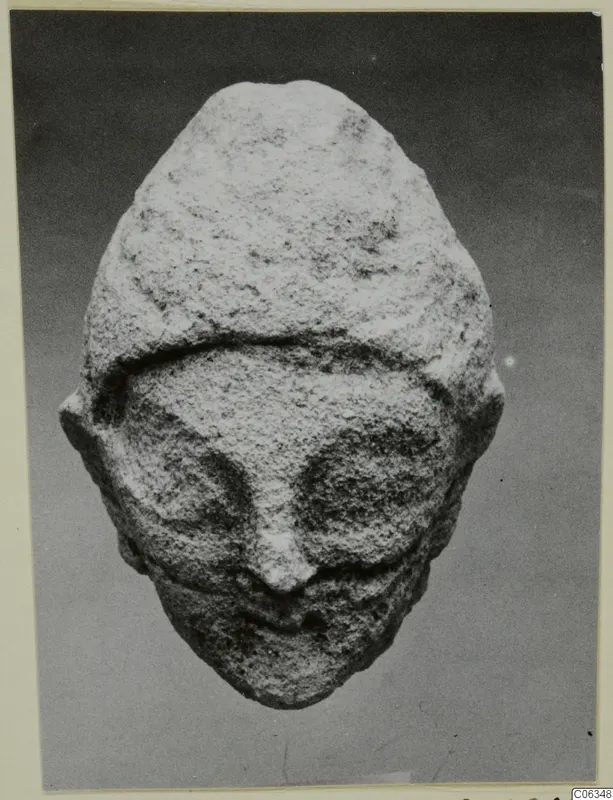 Limestone head from a statuette from Kition, frontal view with prominent nose and deep eye hollows, against a grey backdrop.