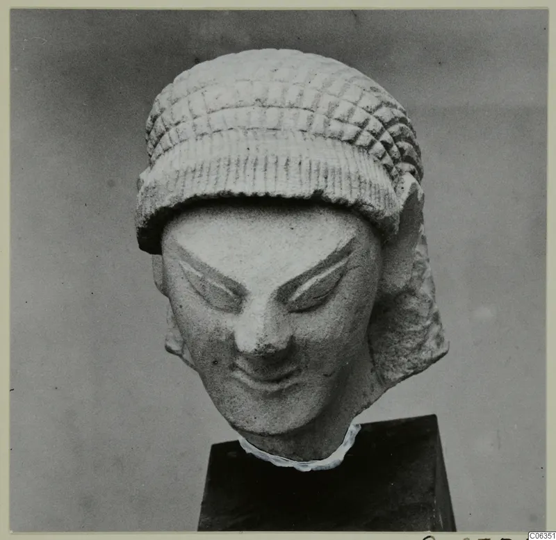 Limestone head statuette from Kition, with a patterned cap and a slight smile, shown on a dark pedestal