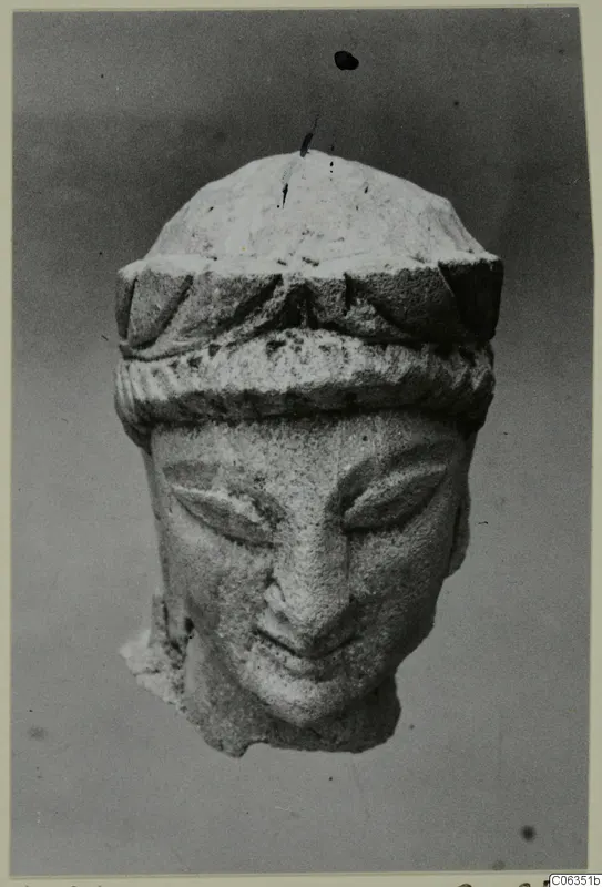 Carved limestone head with a banded headdress and downcast eyes, photographed against a grey background.