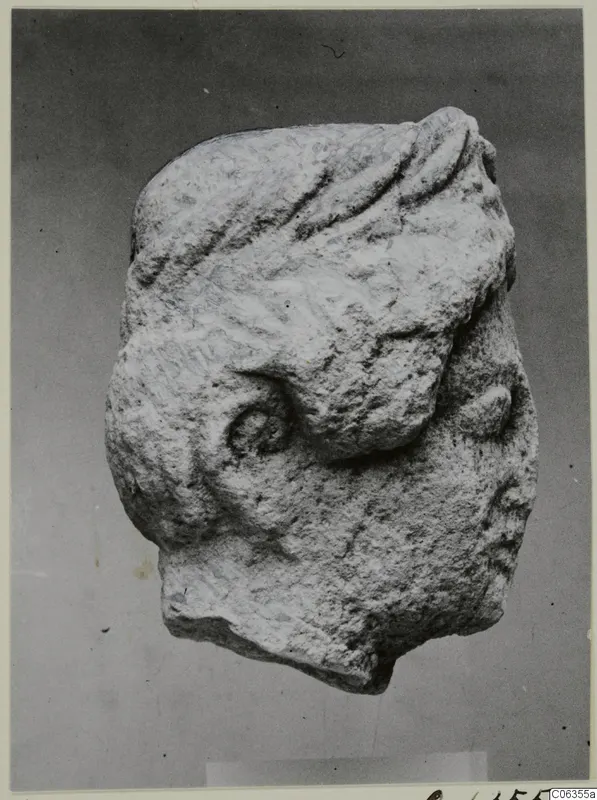 Limestone head fragment with grooved wavy hair and a spiral curl, shown against a plain grey backdrop.