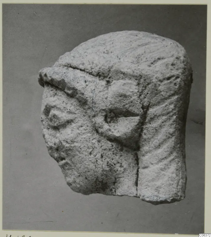 Limestone head fragment from Vouni with carved eyes and grooved hair, shown against a grey backdrop