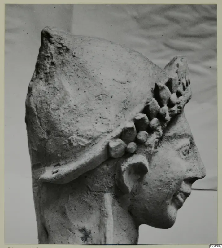 Terracotta head of a statue in profile facing right, with a helmet-like headdress studded with rounded knobs