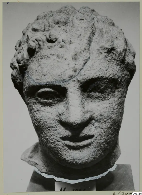 Stone head with curly hair and a crack across the forehead, shown on a dark block base at Mersinaki