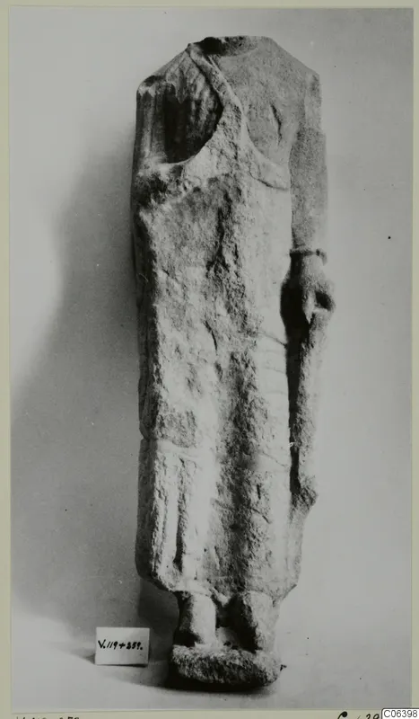 Limestone statue of a standing figure in a long garment, head and shoulders broken away, from Vouni, against a plain grey background.