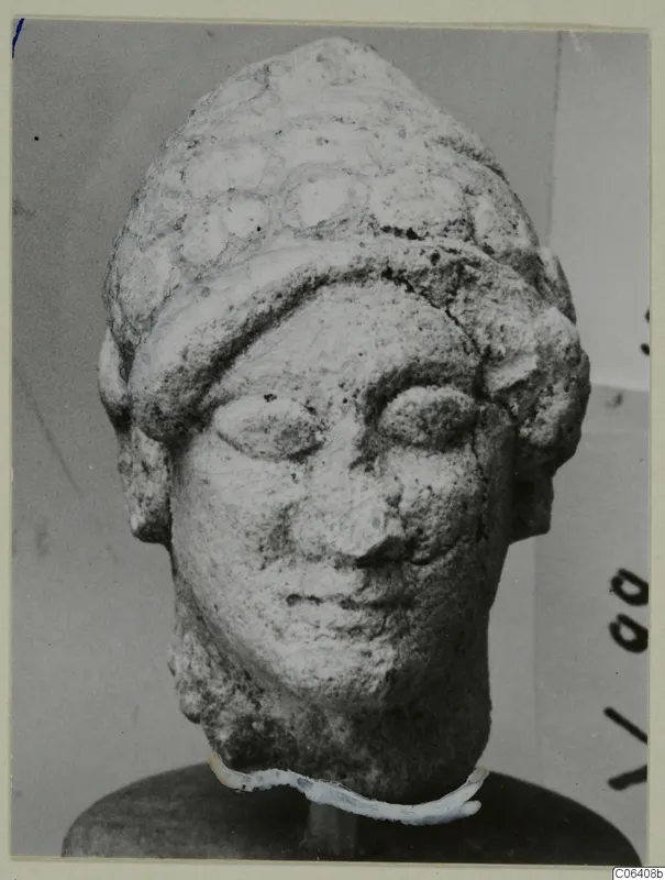 Limestone head from Vouni with a layered headdress, shown front-on on a round wooden stand.