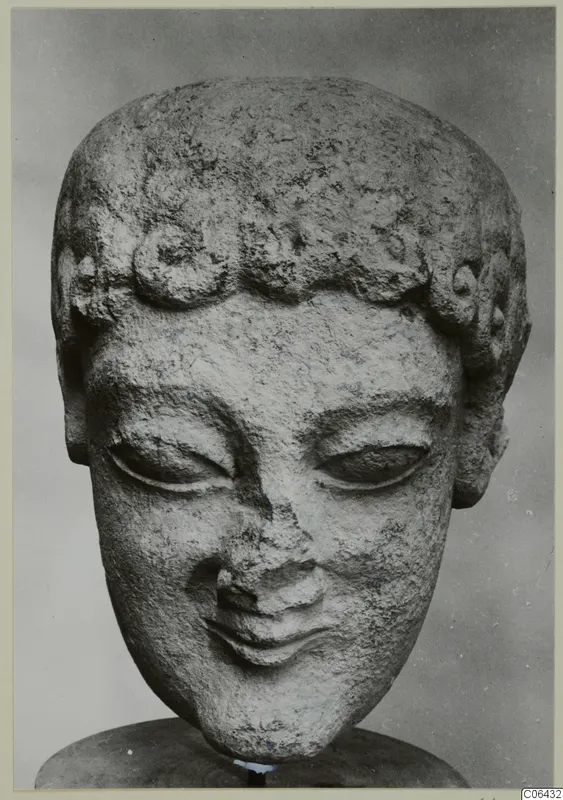 Limestone head of a statue with curled hair and downcast eyes, shown on a dark stand against a grey backdrop.