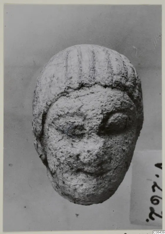Small limestone head with incised lines on the hair, photographed from the front against a grey backdrop.