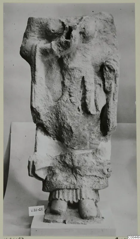 Limestone fragment of a standing figure with the head missing, set on a plinth in front of a grey backdrop