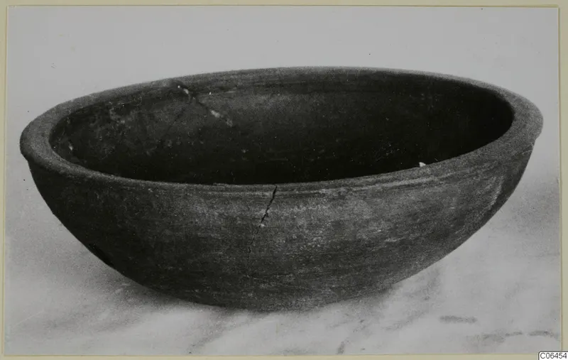 Coarse pottery bowl with a wide, rounded body and dark surface, photographed on a pale surface