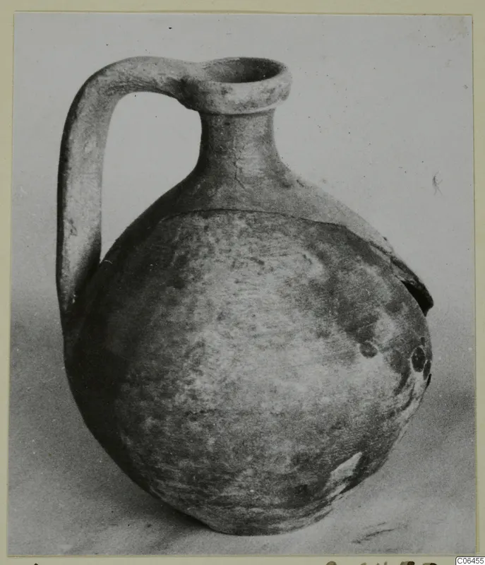 Pottery juglet with a loop handle and round body, photographed against a grey backdrop.