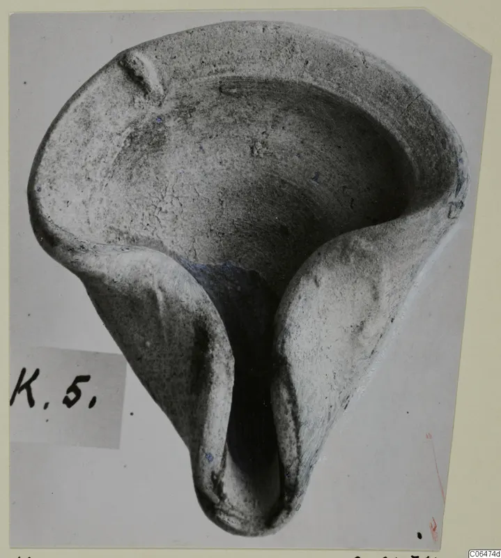 Terracotta lamp with a shallow open bowl and a long spout, photographed from above on a grey mount.