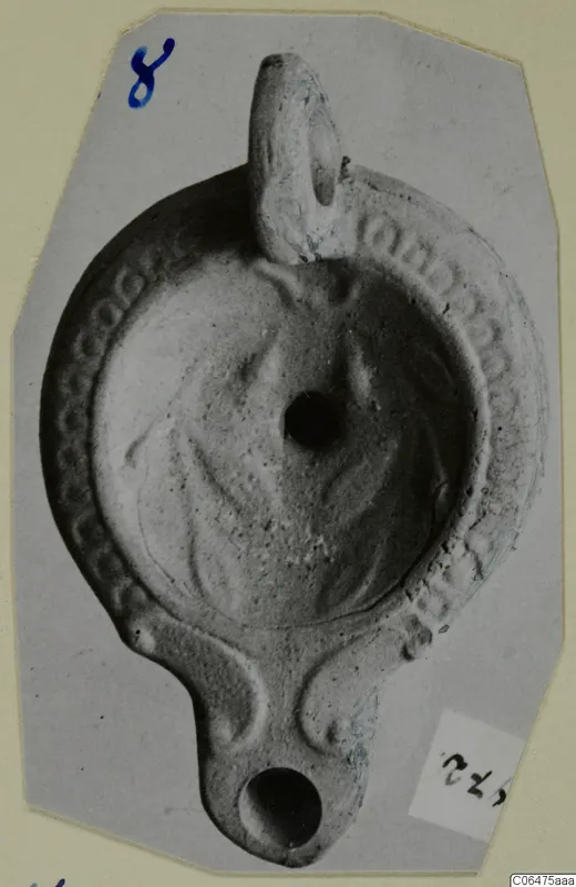 Terracotta oil lamp with a loop handle and a decorated rim, shown on a grey photo mount.