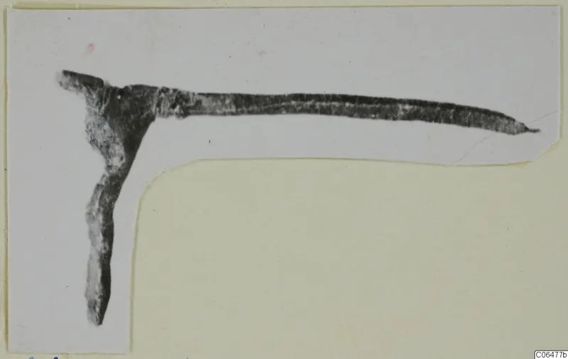 Corroded bronze fragment with a long tapering arm and a shorter arm bent down at a right angle, on a mounted print