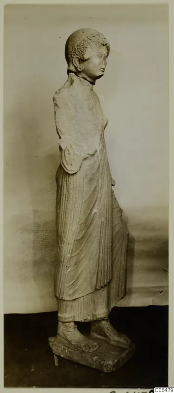 Limestone statue of a standing woman from Vouni, shown in profile in a long pleated garment on a rectangular base.