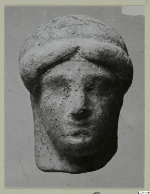 Small terracotta head of a figure with a rounded cap, face frontal, photographed against a grey backdrop