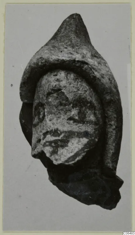 Small terracotta head fragment with a pointed headdress, shown against a plain grey backdrop
