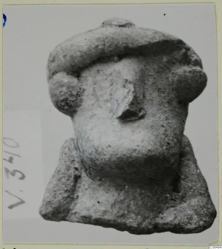Small terracotta figurine fragment with a wide flat-topped headdress and projecting side knobs, photographed on a mounted card.