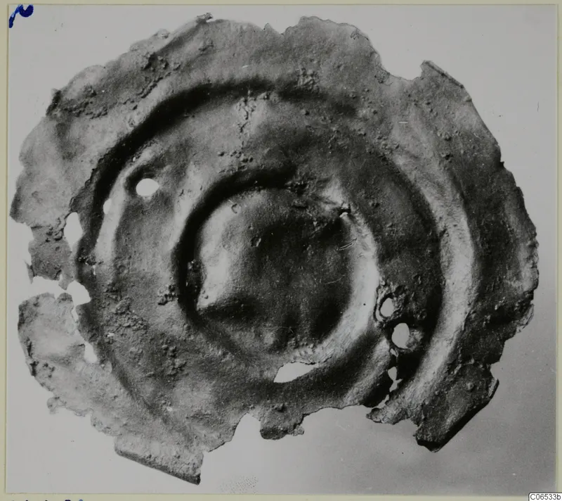 Corroded bronze disc fragment with concentric raised rings and two small holes, from Vouni.
