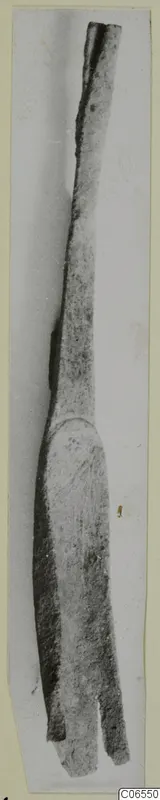 Long bronze implement with a slender shaft and a flattened, split end, shown on a card mount