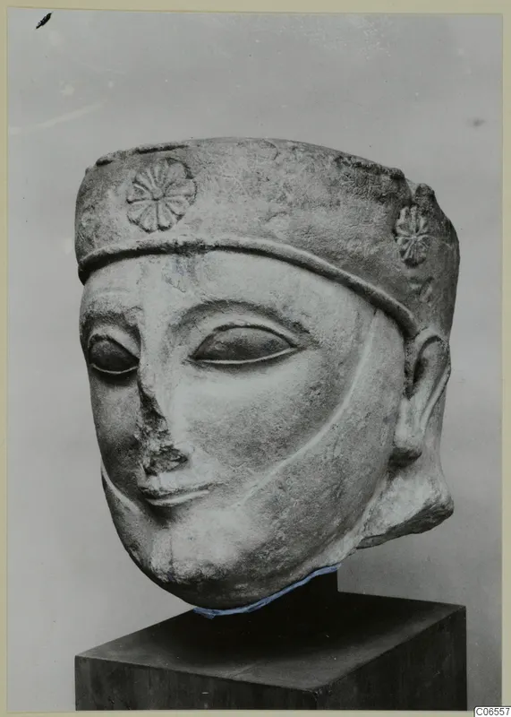 Limestone head sculpture with a tall cylindrical headdress carved with rosettes, shown on a dark pedestal