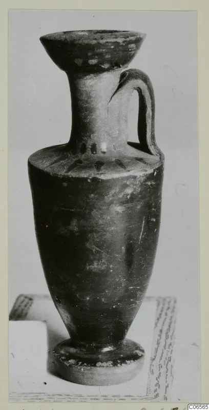 Black glazed ware jug with a flaring rim, a single handle and a ring foot, photographed on a sheet of paper.