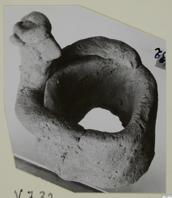 Rough grey clay object with a thick ring-shaped body and a head-like knob on a raised arm, shown on a card mount