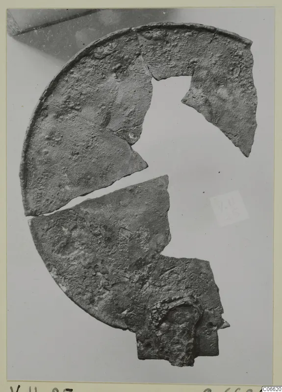 Fragments of a corroded metal disc with a raised rim, from Vouni T. 1-16, shown on a pale backdrop