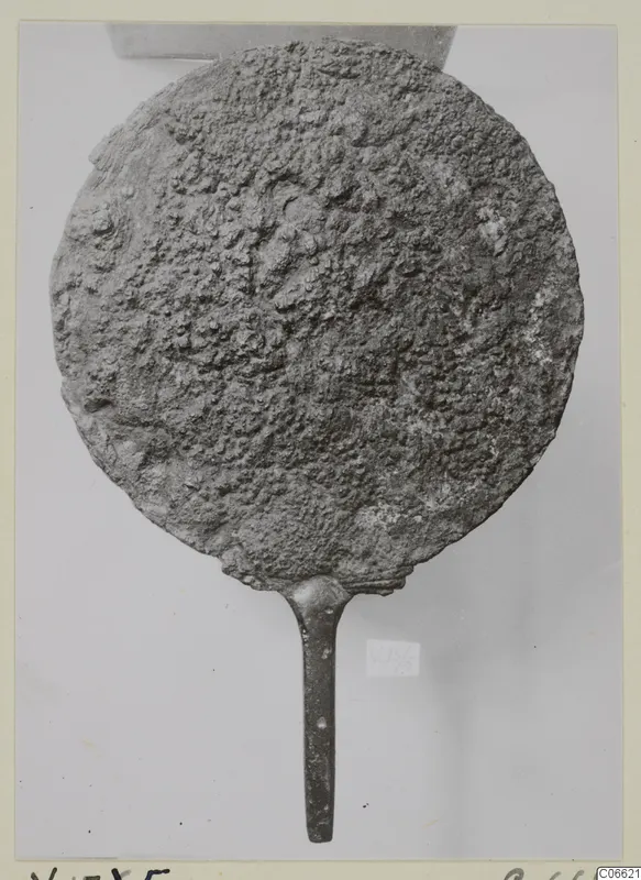 Metal mirror from Vouni: a round disc with a long handle, its surface heavily encrusted with corrosion.