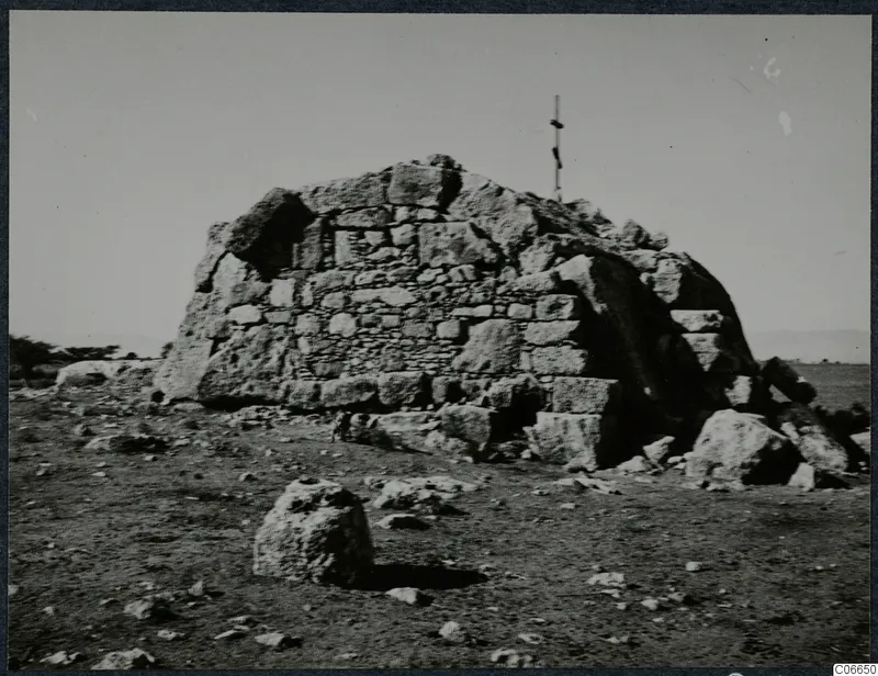 Built tomb of large dressed stone blocks with a pole on top, standing on open, stony ground