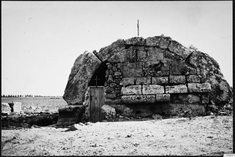 Built stone tomb with a dark doorway and a wooden door marked with a cross, set on bare sand.
