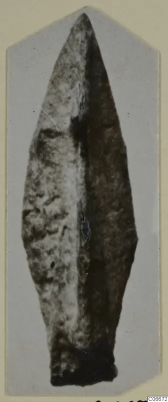 Bronze arrowhead from Sol. 12, a long leaf-shaped point with a central ridge, photographed on a card mount