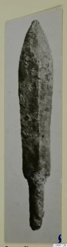 Bronze dagger blade with a pointed tip and short tang, photographed against a grey background.
