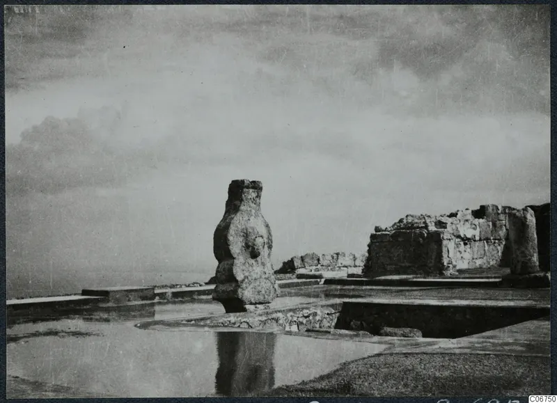 Tall worked stone pillar standing among ruined walls and paved slabs at Vouni palace, with a pale horizon behind.