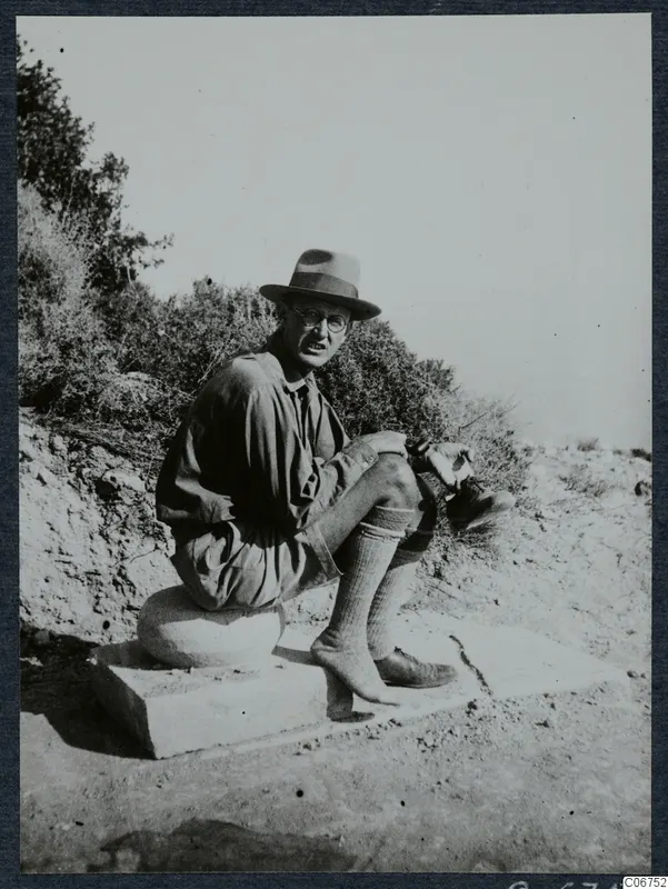 Einar Gjerstad seated on cut stone blocks at Loures, Vouni, with bushes and a bank of soil behind him.