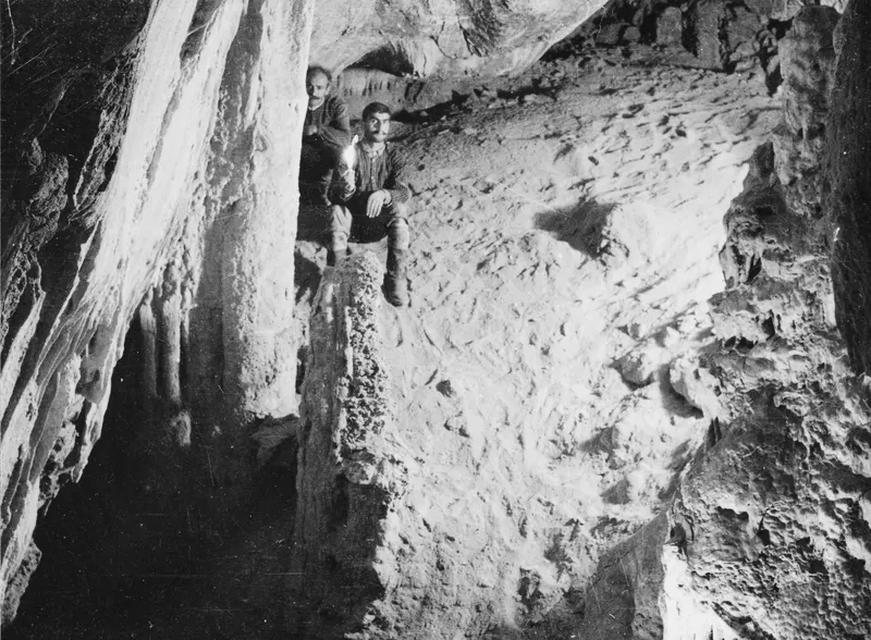 Two men in a cave at Fylleri, Kythrea, one seated on a rock ledge and the other standing behind a column.
