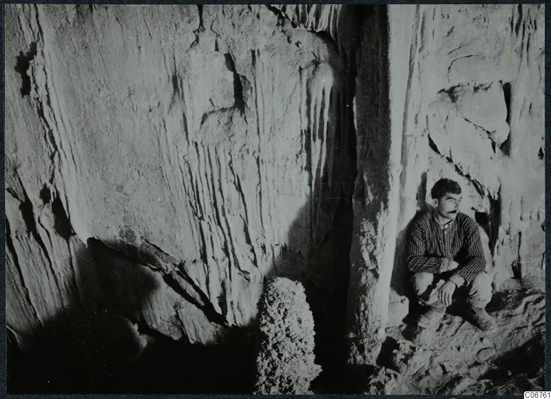 Man crouching in a cave at Kythrea, with rock walls streaked by vertical marks behind him