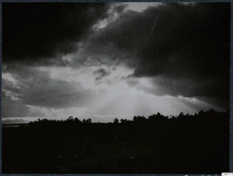 Sunbeams breaking through dark clouds above a line of trees on a flat plain, with faint mountains on the left horizon.