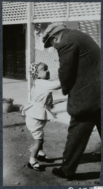 A man in a flat cap and dark coat bends down to play with a small child in a courtyard in Nicosia
