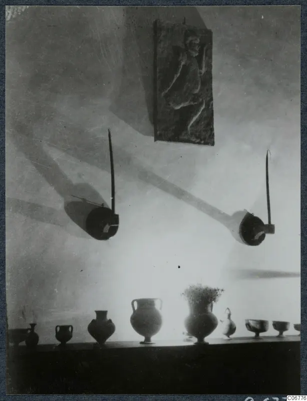Row of vessels on a shelf in a hall, one holding a sprig of flowers, with a relief plaque and two wall-mounted objects above