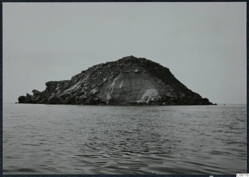 Rocky island rising from the sea under a pale sky, with a steep pale face on its right side