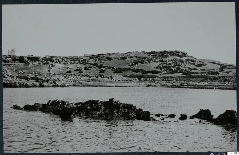 Rocky coastline with low scrubby hills, a small white building on the left, and dark rocks in the sea